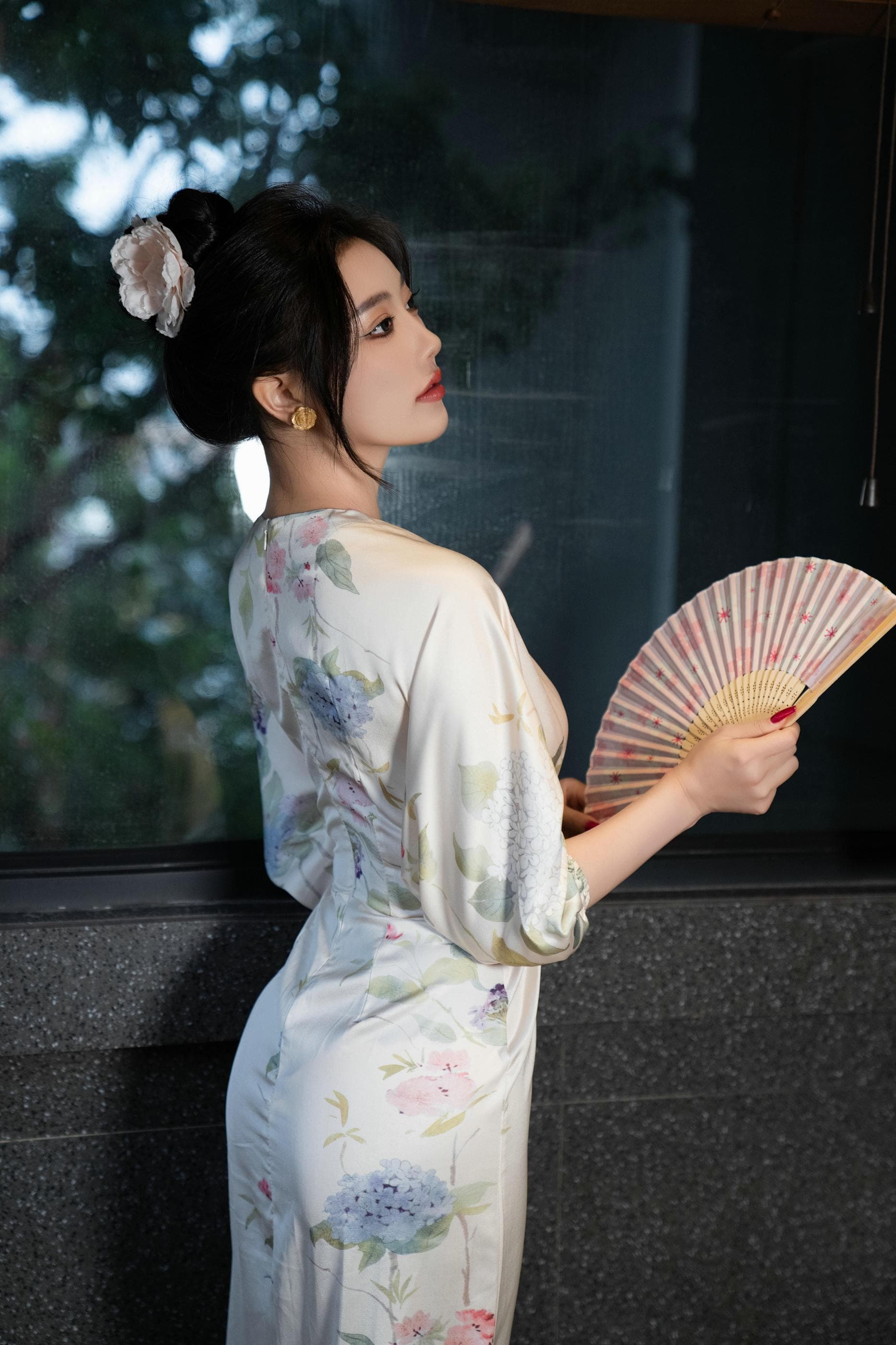 Elegant woman in a floral dress holding a folding fan, standing near a large window.